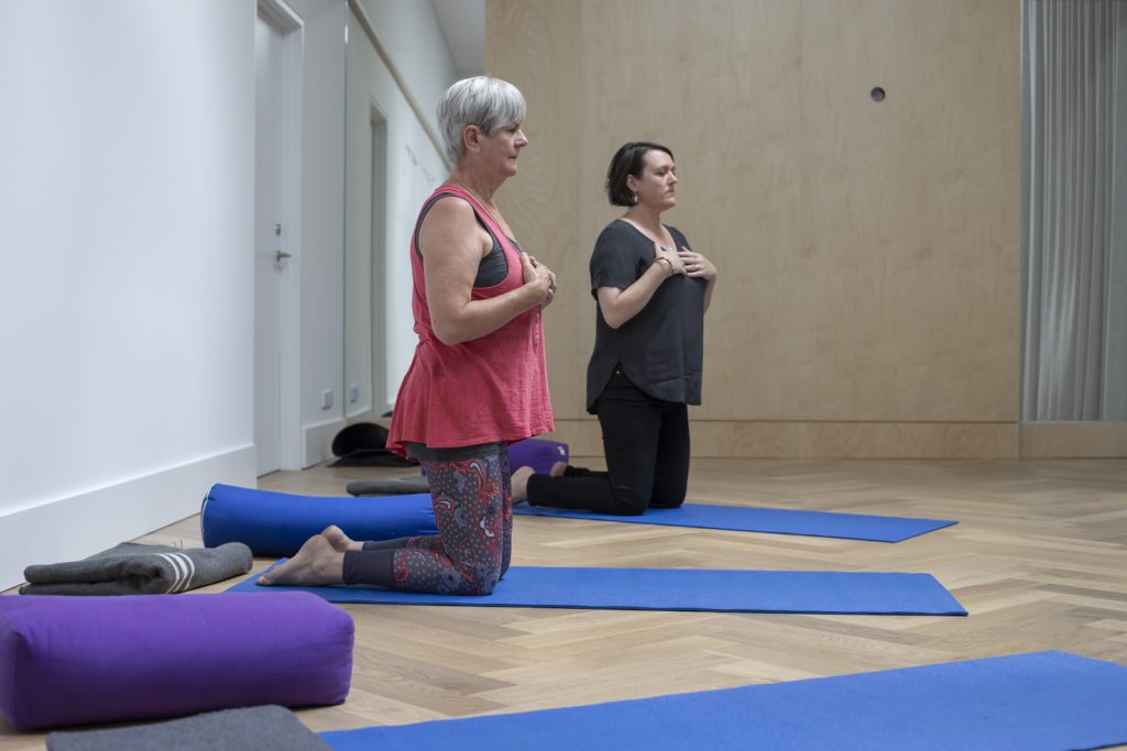 Radha and student, on knees on yoga mats with hands on the chest and eyes closed doing a yoga therapy session inside the yoga studio